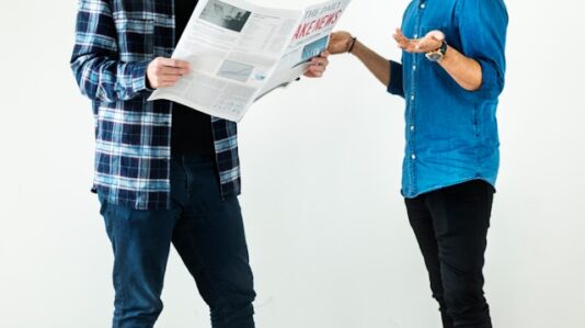 two men standing while one is reading a newspaper